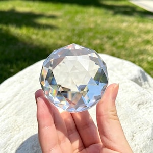 Può includere: Una sfera di cristallo trasparente e sfaccettata tenuta in mano. La sfera è sferica con molte superfici triangolari piatte che riflettono la luce, creando un effetto arcobaleno. Lo sfondo è sfocato con erba verde e luce solare.