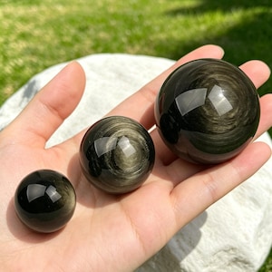 Peut inclure: Trois sphères d'obsidienne polie de tailles différentes sont présentées. Les sphères ont une surface sombre et réfléchissante avec des inclusions dorées tourbillonnantes. La plus petite sphère mesure environ 2.5 cm de diamètre, la moyenne environ 5 cm et la plus grande environ 7.5 cm.