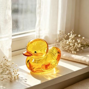 Könnte beinhalten: Eine durchscheinende, bernsteinfarbene Glasentenfigur mit einem glatten, abgerundeten Körper, einem ausgeprägten Schnabel und kleinen dunklen Augen. Die Ente ist aus klarem gelbem Glas gefertigt, das das Licht einfängt und hell reflektiert.