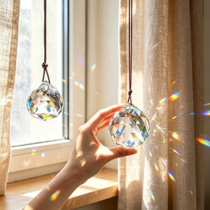 May include: Two clear, multifaceted crystal spheres suspended by thin brown cords. Each glass prism features geometric triangular facets that refract sunlight, casting vibrant rainbow light patterns onto the surrounding area near a window.