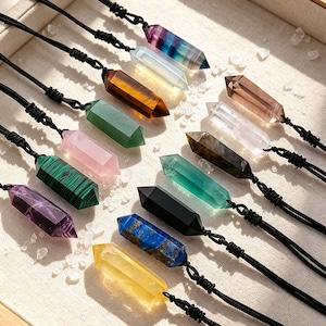 May include: A collection of twelve hexagonal crystal point pendants on black cord necklaces. The stones feature various colors including purple, green, pink, blue, yellow, black, brown, and clear, arranged in rows on a light fabric surface.