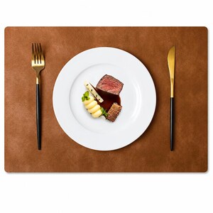 May include: A rectangular brown leather placemat featuring a white ceramic plate centered in the middle. The plate holds a small portion of cooked meat, a swirl of mashed potatoes, and a garnish. A gold and black fork and knife are placed on the sides.