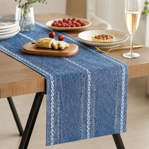 May include: A blue fabric table runner featuring vertical white woven patterns and textured stripes. The runner is laid across a wooden table, serving as a base for a wooden serving board with sliced bread, plates of fruit, and a glass of white wine.