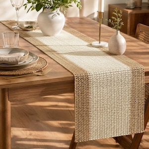 May include: A rectangular table runner featuring a woven texture with a wide cream-colored center panel flanked by two brown side borders. The runner is made of a durable, textured material and displays a consistent, repeating geometric weave pattern.