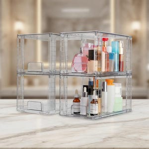 May include: Clear, stackable acrylic storage containers filled with various cosmetic products. The containers are arranged in a two-by-two configuration, showcasing perfumes, lotions, and other beauty items. The containers sit on a marble-patterned surface.