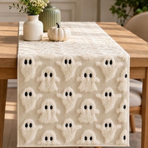 May include: A cream-colored table runner featuring a repeating pattern of raised, textured white ghosts with black embroidered eyes. The runner is laid flat on a wooden table, showcasing a soft, plush fabric construction with finished edges.