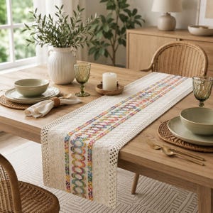 May include: A cream-colored table runner featuring a central band of colorful, embroidered oval patterns in shades of blue, pink, yellow, and green. The runner has a textured, woven fabric base with a decorative crocheted edge, laid flat on a wooden table.