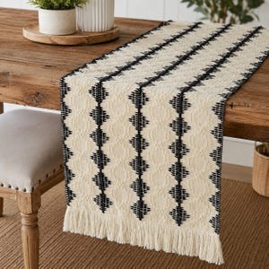 May include: A cream-colored table runner featuring a repeating black geometric diamond pattern and vertical stripes. The runner has a textured woven surface and is finished with long, straight fringe along the bottom edge of the fabric.