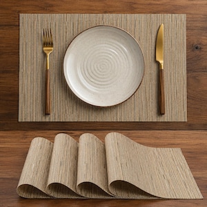 Puede incluir: Un juego de manteles individuales rectangulares tejidos en color beige neutro con acabado texturizado. Un mantel está extendido con un plato de cerámica blanca con patrón en espiral, un tenedor dorado y un cuchillo dorado con mango de madera.