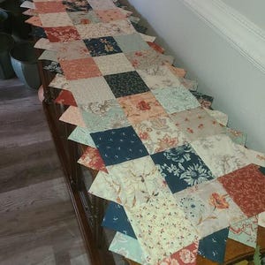 May include: A patchwork table runner with a scalloped edge. The runner features a variety of floral and patterned fabrics in shades of red, blue, cream, and peach. The runner is placed on a dark wood table.