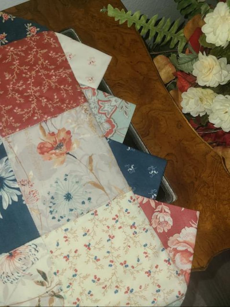 May include: A patchwork of fabric squares in various floral patterns and colors, including red, white, blue, and cream. The fabrics are arranged in a geometric design, with visible stitching. The image is taken from an elevated angle, showing the fabric arrangement in a tray.