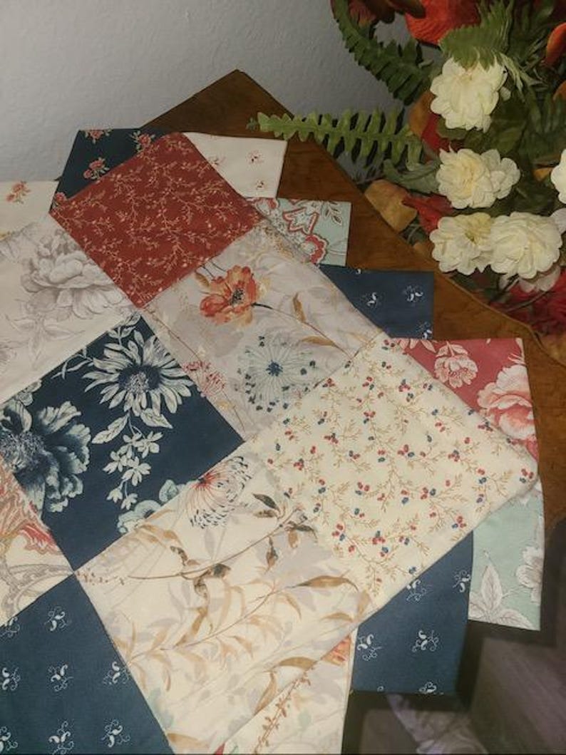 May include: A patchwork quilt with a variety of floral patterns in shades of blue, red, cream, and gray. The quilt is made of square fabric pieces arranged in a diagonal pattern. The quilt is placed on a wooden surface.