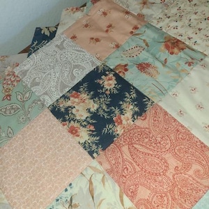 May include: A patchwork quilt with a variety of floral and paisley patterns in shades of blue, peach, and cream. The quilt is made of square fabric pieces sewn together, creating a textured surface.