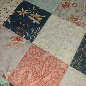 May include: Close-up of a patchwork quilt with various floral and paisley patterns. The quilt features diamond-shaped patches in shades of blue, cream, peach, and gray. Some patches have large floral designs, while others have delicate, repeating patterns.