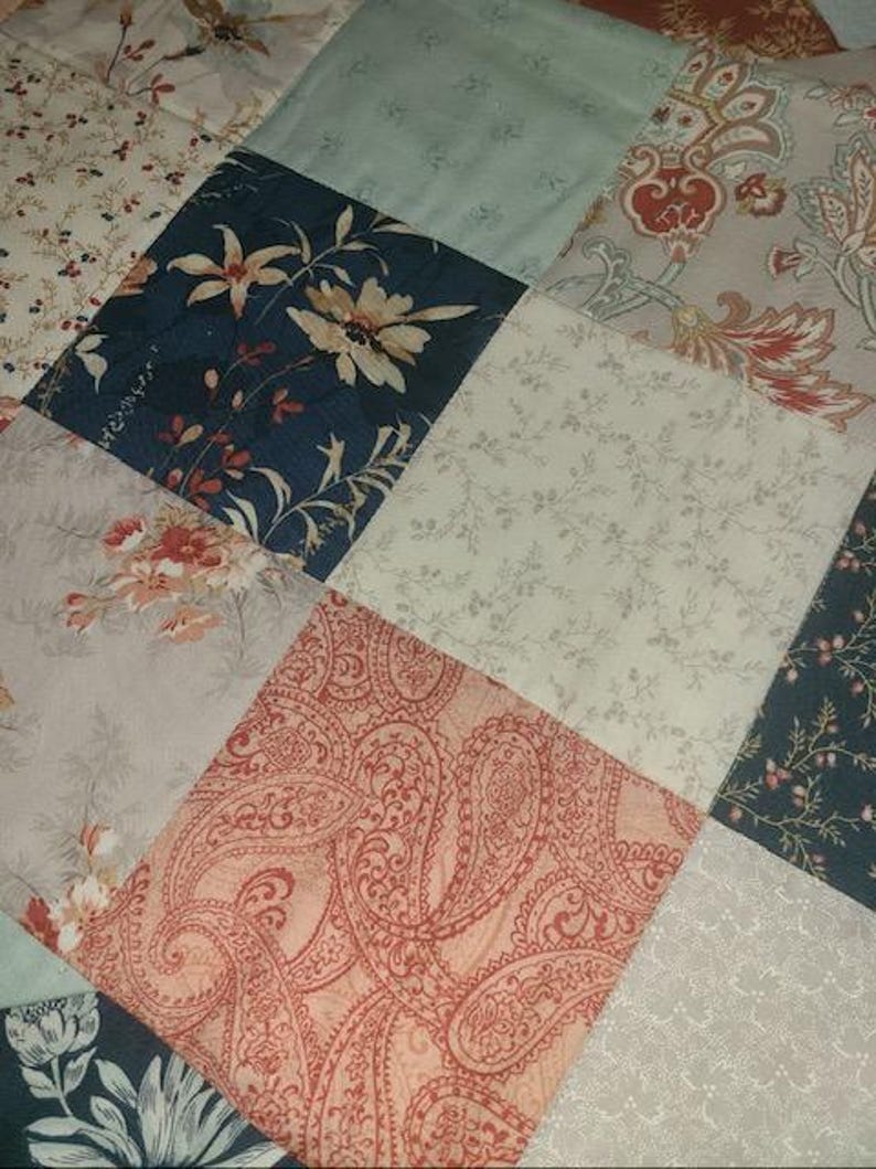 May include: Close-up of a patchwork quilt with various floral and paisley patterns. The quilt features diamond-shaped patches in shades of blue, cream, peach, and gray. Some patches have large floral designs, while others have delicate, repeating patterns.