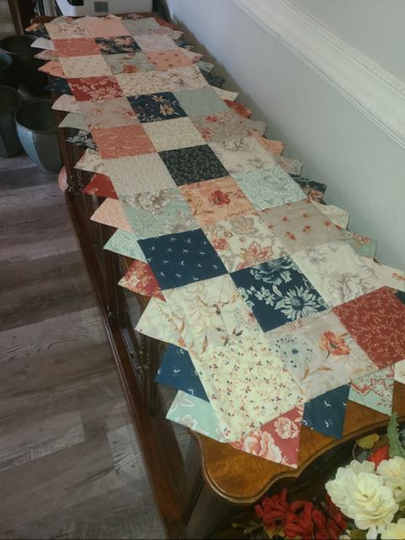 May include: A patchwork table runner with a scalloped edge. The runner features a variety of floral and patterned fabrics in shades of red, blue, cream, and peach. The runner is placed on a dark wood table.