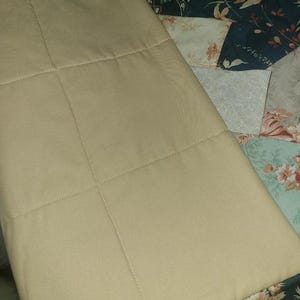 May include: A tan, quilted fabric item with a grid pattern, possibly a pillow or a part of a larger textile. The item is set against a backdrop of a patchwork quilt with floral and geometric patterns in various colors.