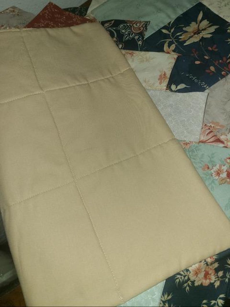 May include: A tan, quilted fabric item with a grid pattern, possibly a pillow or a part of a larger textile. The item is set against a backdrop of a patchwork quilt with floral and geometric patterns in various colors.