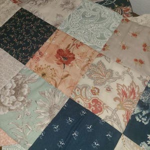 May include: A patchwork quilt featuring a variety of floral and paisley patterns in shades of pink, green, blue, and beige. The quilt is made of square fabric pieces sewn together, creating a textured surface. The overall design is a mix of vintage and modern styles.