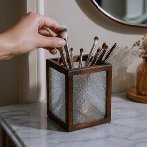 Peut inclure: Un porte-pinceaux de maquillage carré doté d'un cadre en bois brun foncé et de panneaux en verre texturé avec un motif floral répétitif. Le récipient contient plusieurs pinceaux de maquillage aux manches argentés et aux poils variés.