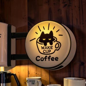 May include: A circular wall-mounted light box sign featuring a black cat graphic inside a coffee cup with the text WAKE CUP and Coffee printed below. The sign emits a warm yellow glow and is mounted on a dark wooden surface with coffee mugs.