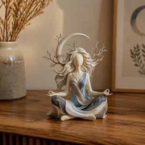 May include: A figurine of a figure in a seated meditation pose with flowing hair, wearing a blue and white draped garment. Behind the figure is a crescent moon shape and bare, branching twigs. The sculpture features muted blue, grey, and cream tones.