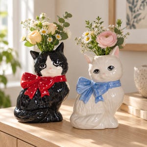 Puede incluir: Dos jarrones de cerámica con forma de gato que contienen arreglos florales. Un gato es negro con un lazo rojo de lunares y el otro es blanco con un lazo azul de lunares. Ambos tienen detalles faciales pintados, acabados brillantes y aberturas.