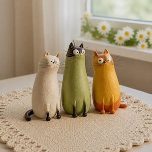 Peut inclure: Trois figurines de chats aux corps allongés et aux grands yeux. Le chat de gauche est crème avec une queue noire, celui du centre est vert olive avec une tête noire, et celui de droite est orange avec le bas du corps plus clair et une petite queue.
