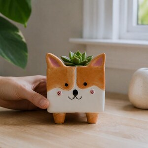 Pode incluir: Um vaso de cerâmica quadrado em forma de cão corgi com esmalte laranja e branco, orelhas internas cor-de-rosa e um rosto pintado. O vaso possui quatro pequenos pés cor de laranja e contém uma pequena planta suculenta verde no topo.