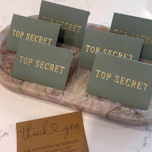 May include: Sage green cards with the words "TOP SECRET" in gold lettering are arranged on a decorative tray. A brown card with the words "thank you" in cursive is also visible.