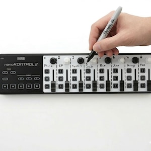 Może przedstawiać: Czarny kontroler MIDI Korg nanoKONTROL2 z białymi i czarnymi suwakami i przyciskami. Ręka trzymająca marker Sharpie pisze na kontrolerze. Kontroler ma etykiety dla Piano, EP, Synth, Sub, Bass, Arp, Strings i PAD.
