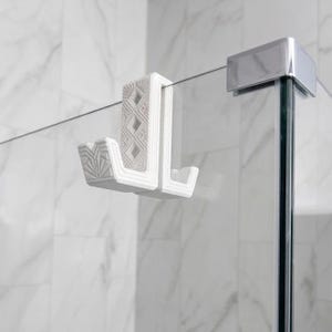 May include: A white shower door hook with an Art Deco design. The hook has a geometric pattern and is made of a solid material. It is designed to hang over a glass shower door.