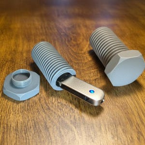 Puede incluir: Un pendrive gris en forma de perno con un conector USB plateado y una luz azul. El pendrive está parcialmente insertado en un contenedor gris en forma de perno, con una cabeza de perno a juego y una tuerca de perno separada.