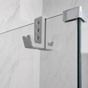 May include: A light gray over-the-glass shower hook featuring a textured geometric pattern on the front and side surfaces. The hook has a U-shaped design that fits over the top edge of a glass shower panel, providing a sturdy mount for towels.
