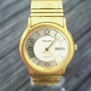 Peut inclure: Une montre Gruen Quartz dorée avec un cadran argenté et blanc. La montre affiche la date et possède des aiguilles dorées. Le bracelet de la montre est également doré. Le cadran affiche les chiffres 12, 1, 2, 5, 6, 9, 10 et 11.