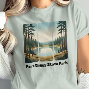 May include: A pale green t-shirt with a landscape design showing a lake and trees. The text "Fort Boggy State Park" is below the image. The t-shirt is a casual wear item, suitable for everyday use.