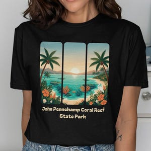 Op de afbeelding: Zwart T-shirt met een schilderachtige illustratie van John Pennekamp Coral Reef State Park. Het ontwerp toont een tropische strandscène met palmbomen, oceaan en kleurrijke bloemen. De tekst "John Pennekamp Coral Reef State Park" staat eronder.