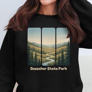 May include: Black sweatshirt with a landscape design depicting a river winding through a valley, framed by trees and mountains. The text "Buescher State Park" is printed below the image.