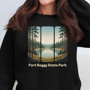 May include: Black sweatshirt with a landscape design depicting a lake, trees, and mountains. The text "Fort Boggy State Park" is printed in white. The design is framed within three vertical rectangles, creating a scenic view.