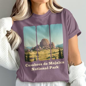 May include: A mauve t-shirt with a landscape illustration of Cumbres de Majalca National Park. The design shows a mountain range, a field, and trees, framed by three vertical blue panels. The text "Cumbres de Majalca National Park" is printed below the image.