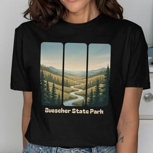May include: Black t-shirt with a landscape design depicting a river, hills, and trees. The text "Buescher State Park" is printed below the image in white. The design is framed within three rectangular panels.