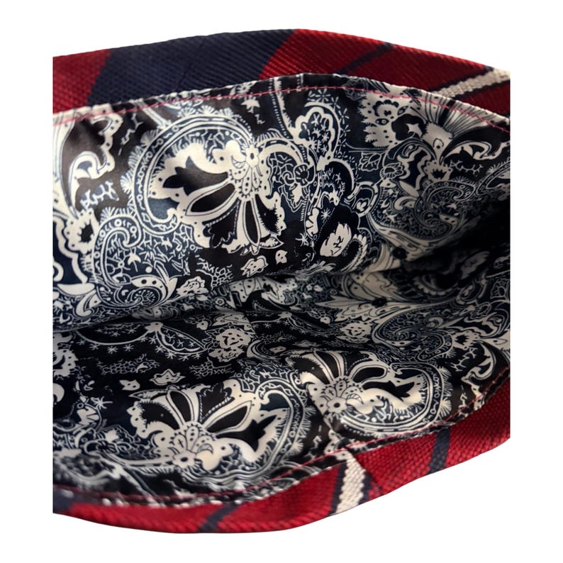 May include: A bag interior featuring a black and white paisley pattern lining. The exterior edge of the bag is constructed from a red and navy blue plaid fabric with a visible woven texture, showing the contrast between the outer and inner layers.