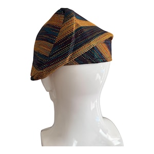 May include: A multi-colored newsboy cap with a geometric pattern. The cap features a combination of gold, teal, and dark purple stripes. The hat is displayed on a white mannequin head, showcasing its design and shape.