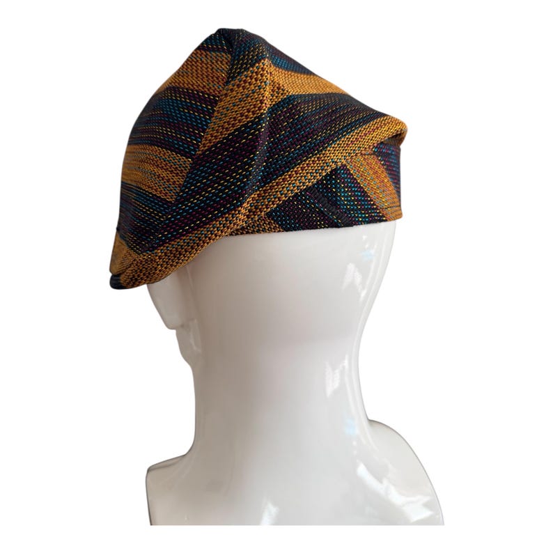 May include: A multi-colored newsboy cap with a geometric pattern. The cap features a combination of gold, teal, and dark purple stripes. The hat is displayed on a white mannequin head, showcasing its design and shape.