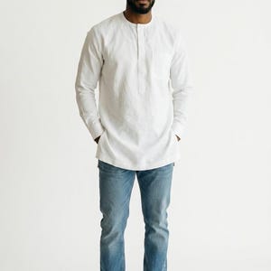 May include: A white long-sleeved tunic shirt with a mandarin collar and a single chest pocket, paired with blue denim jeans and white sneakers. The outfit is completed by a small, pointed cap featuring a maroon, navy, and white striped pattern.