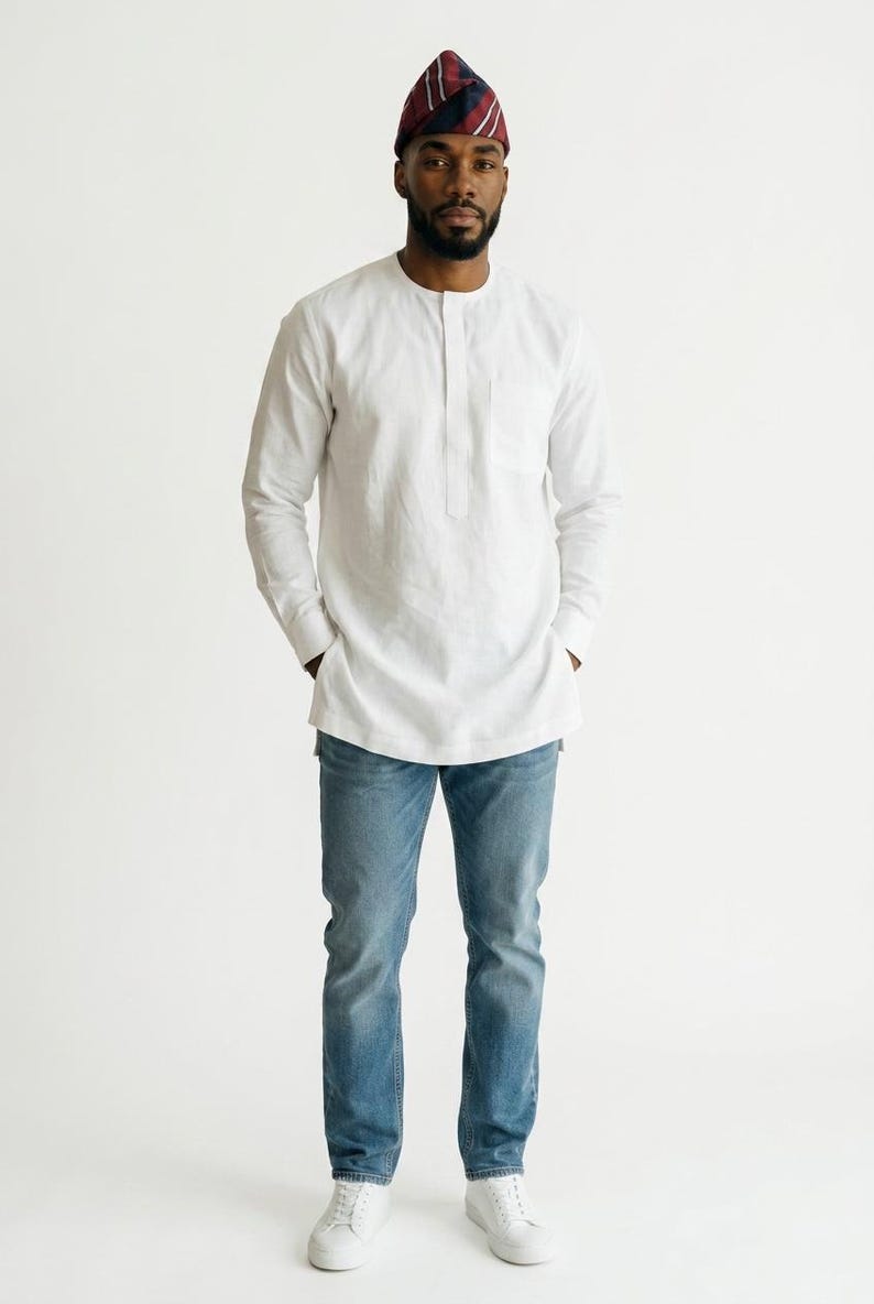 May include: A white long-sleeved tunic shirt with a mandarin collar and a single chest pocket, paired with blue denim jeans and white sneakers. The outfit is completed by a small, pointed cap featuring a maroon, navy, and white striped pattern.