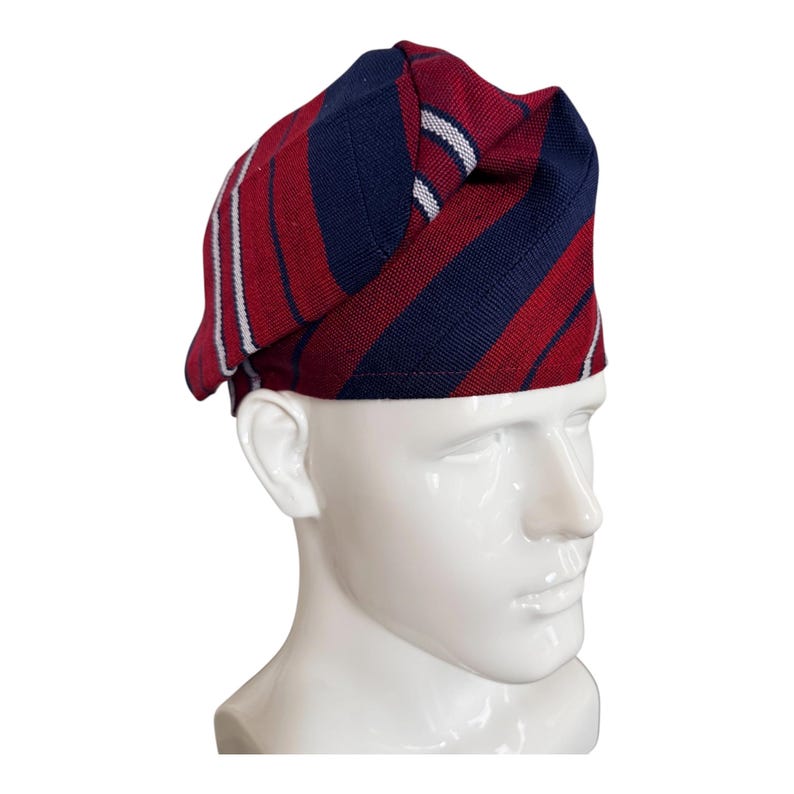 May include: A structured cap featuring a woven pattern of diagonal stripes in deep red and navy blue, accented by thin white lines. The fabric has a textured, knit-like appearance with a folded, layered design that sits atop a white mannequin head.