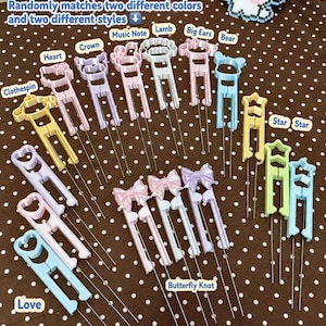 May include: A collection of plastic hair clips in various pastel colors and shapes, including hearts, crowns, music notes, lambs, bears, stars, and bows. Each clip features a long, thin metal prong attachment. Text on the image includes ALL STYLES AVAILABLE DESIGNS, Randomly matches two different colors and two different styles, Random styles, random matches, Multiple options to choose from, and Please leave a note when ordering.