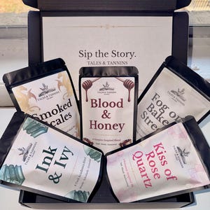 Puede incluir: Una caja negra abierta que contiene cinco bolsas de té con varios sabores. Las bolsas están etiquetadas con nombres como "Blood & Honey" y "Kiss of Rose Quartz". La caja tiene el texto "Sip the Story. TALES & TANNINS."