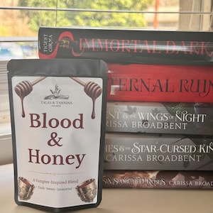May include: A black pouch of "Blood & Honey" tea blend with honey dipper illustrations and text. The pouch is in front of a stack of books with titles like "Immortal Dark" and "Eternal Ruin". The tea is a vampire-inspired blend.